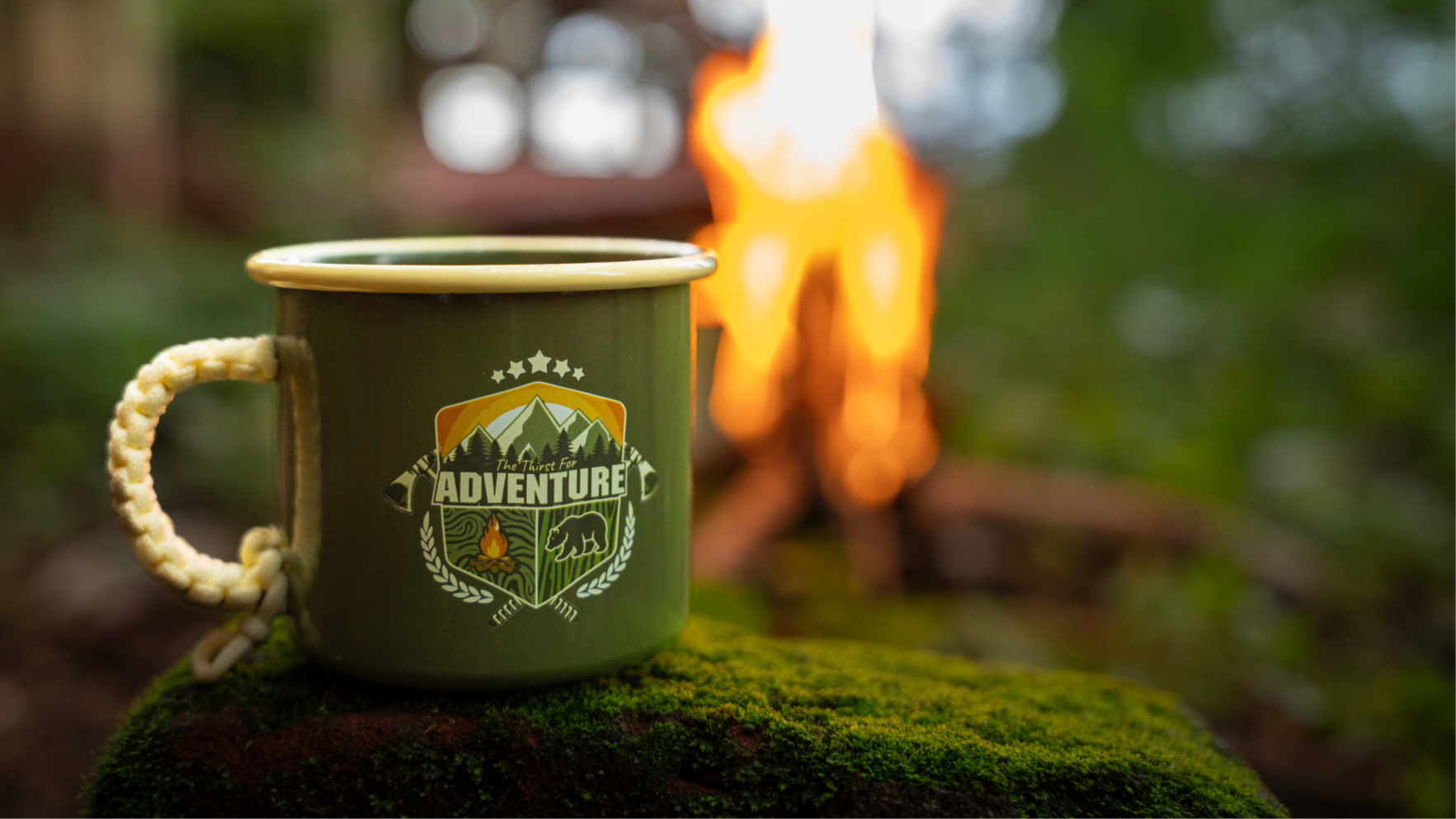 Green mug with 'Adventure' design in front of a campfire in a forest setting